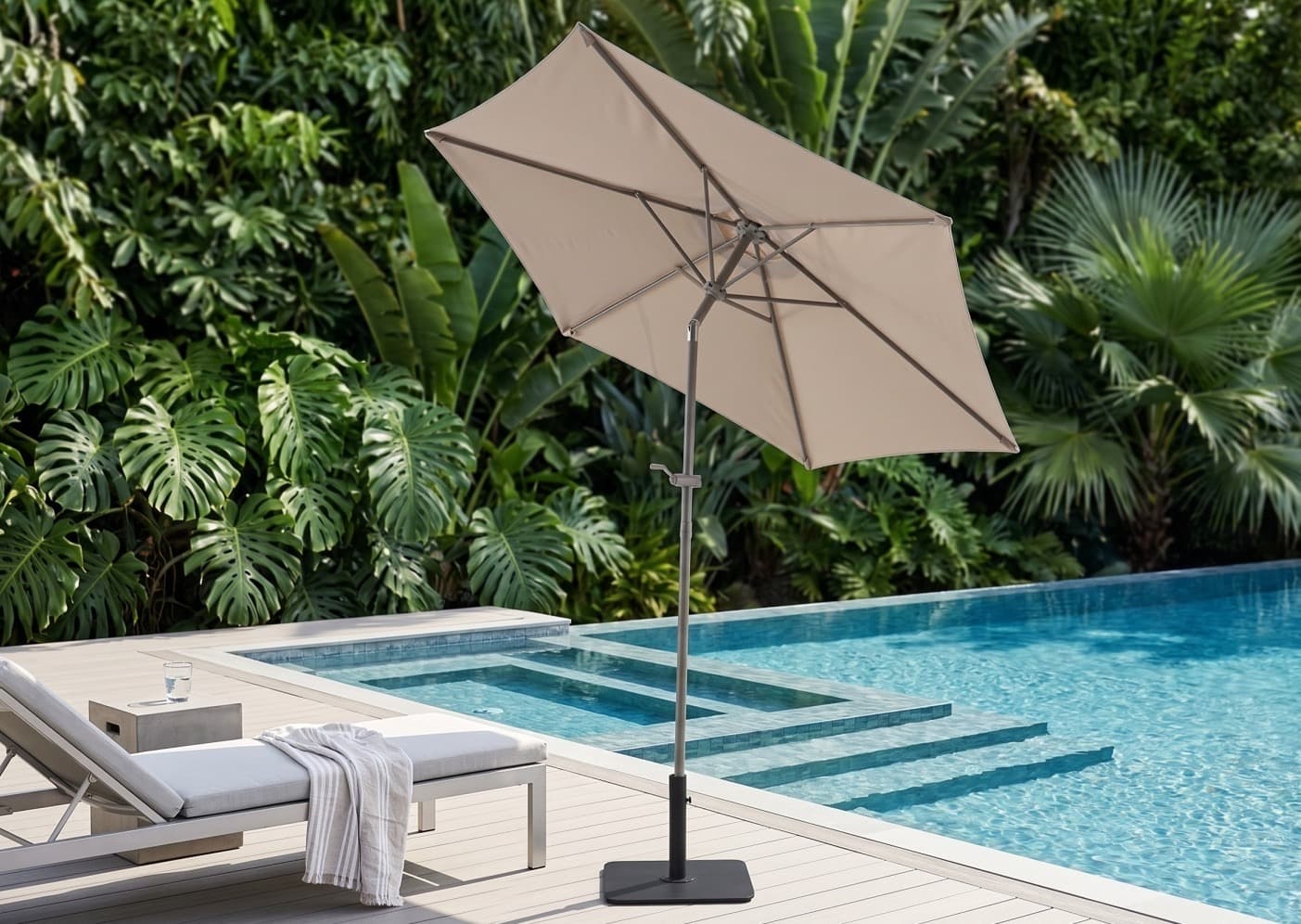 Ombrelone central Brisa em cor areia, com cobertura inclinada, ao lado de um espreguiçadeira à beira da piscina com folhagem tropical ao fundo